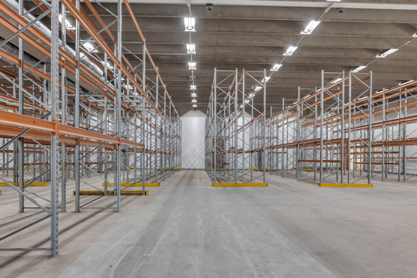 Tyhjä teollisuusvarasto, jossa on useita rivejä metallisia varastohyllyjä ja betonilattia kirkkaiden kattovalojen alla.