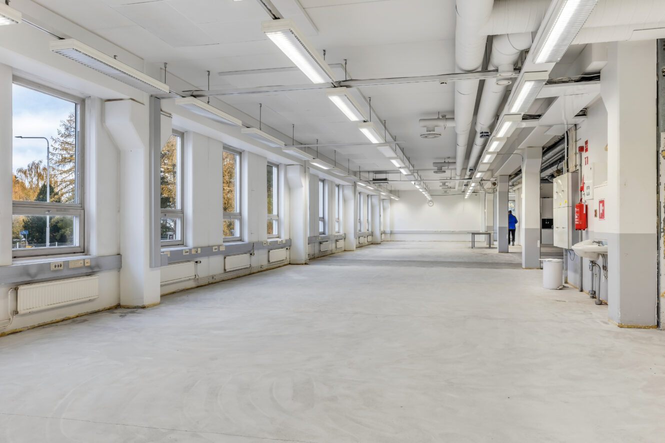 Suuri, tyhjä teollisuushuone, jossa on valkoiset seinät, korkeat ikkunat, katossa paljaat putket ja minimaalinen kalustus.