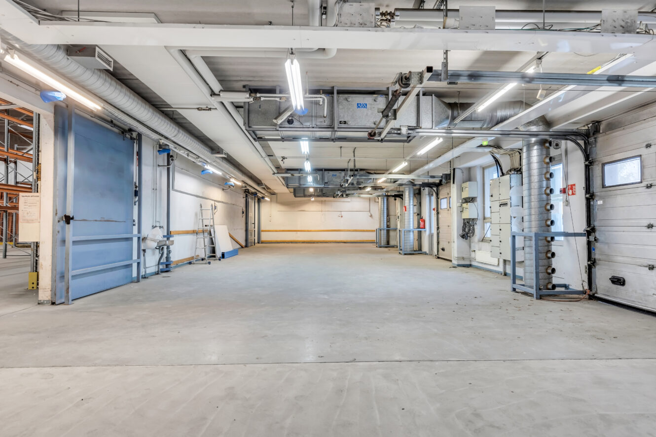 Tyhjä teollisuusvaraston sisätila, jossa on betonilattia, ylävalot, suuret autotallin ovet ja näkyvä kanavisto katossa.