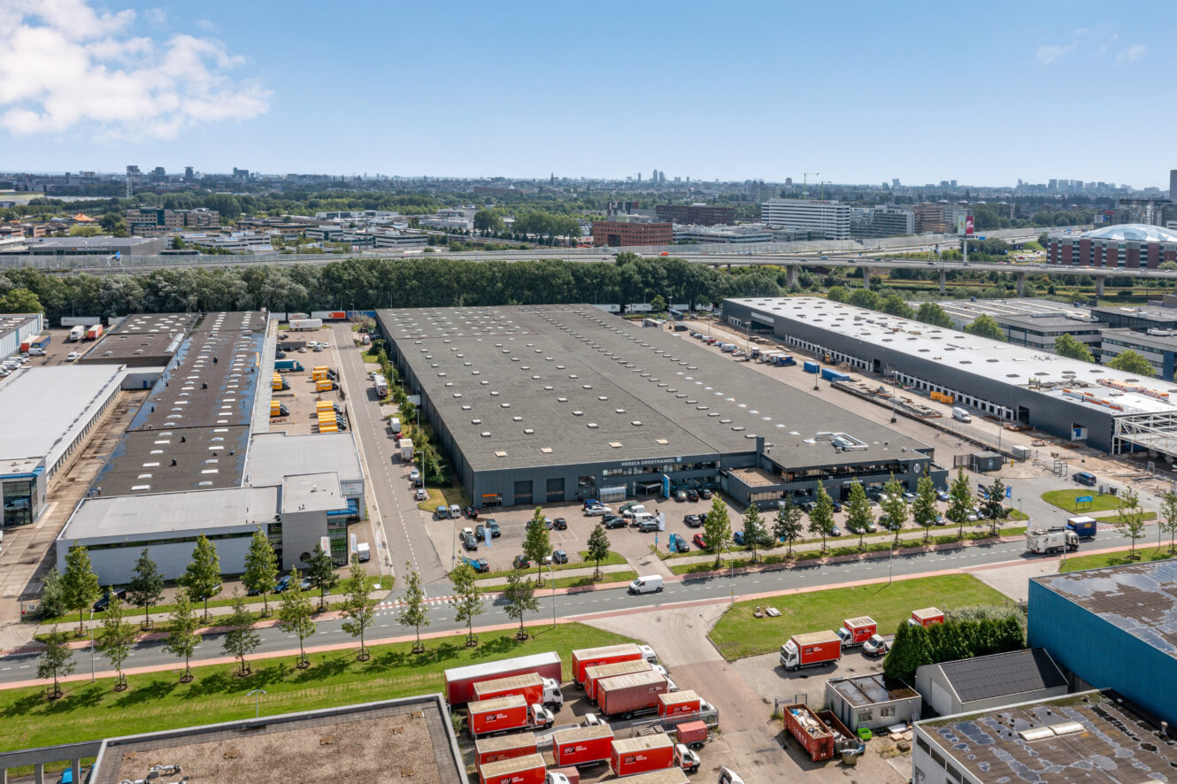 Luchtfoto van een industriegebied met grote magazijnen, parkeerplaatsen, bomen en verschillende rood-witte bestelwagens op wegen en aangrenzende percelen onder een heldere hemel.