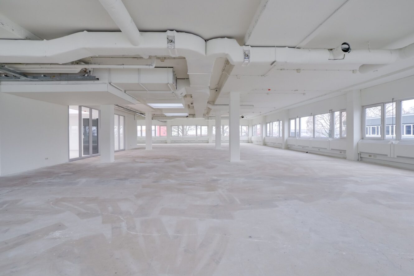 Lege, onafgewerkte kantoorruimte met grote ramen, zichtbare plafondpijpen, witte muren en een kale betonnen vloer. Natuurlijk licht valt binnen via ramen aan twee kanten.