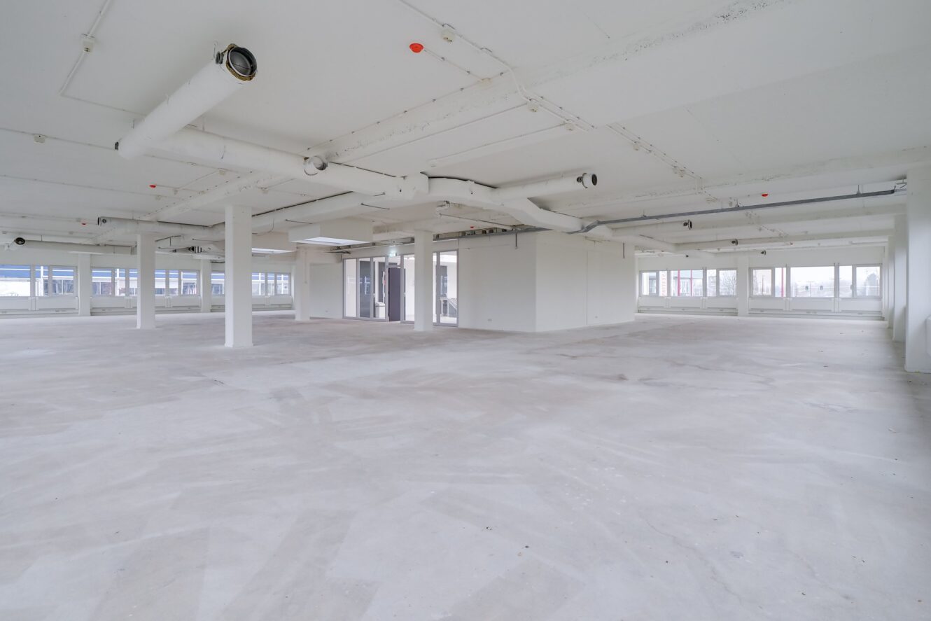 Ruim, leeg commercieel interieur met witte muren, zichtbare buizen, grote ramen en betonnen vloeren.