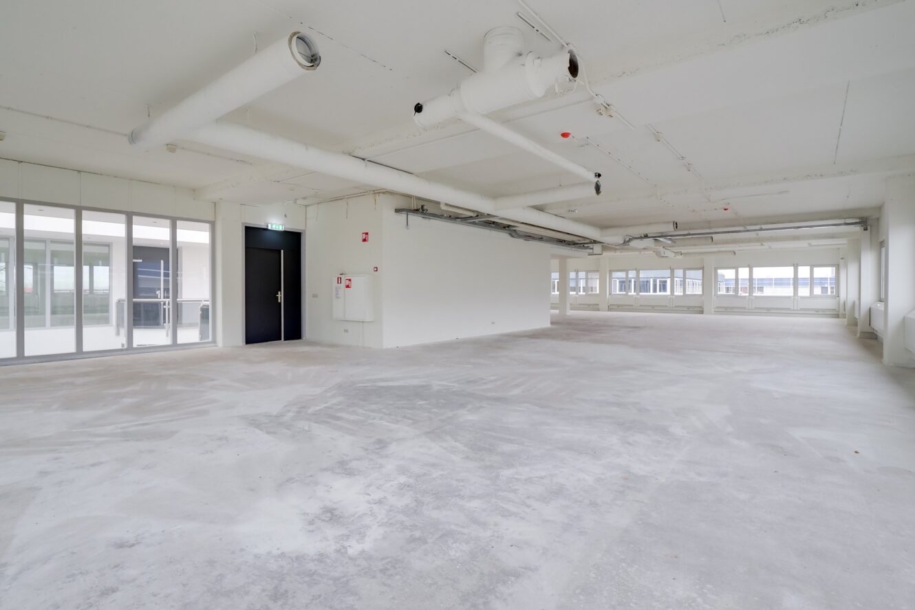 Een lege, onafgewerkte kantoorruimte met kale betonnen vloeren, witte muren, zichtbare buizen aan het plafond en grote ramen die natuurlijk licht binnenlaten.