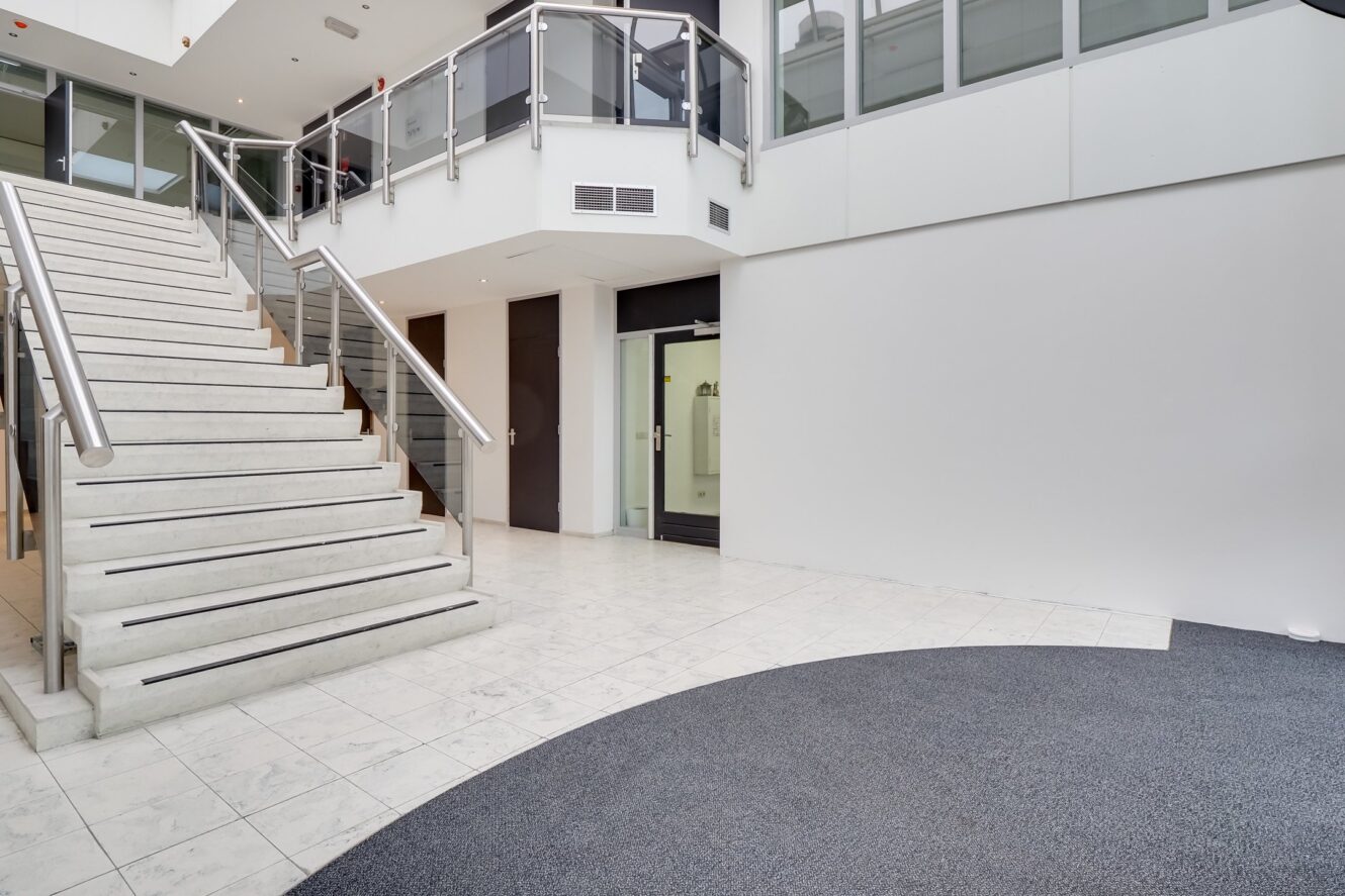 Modern interieur van een gebouw met een witte trap, glazen leuningen, tegelvloer en grote ramen op de bovenverdieping. Op de voorgrond is een ruimte met tapijt zichtbaar.