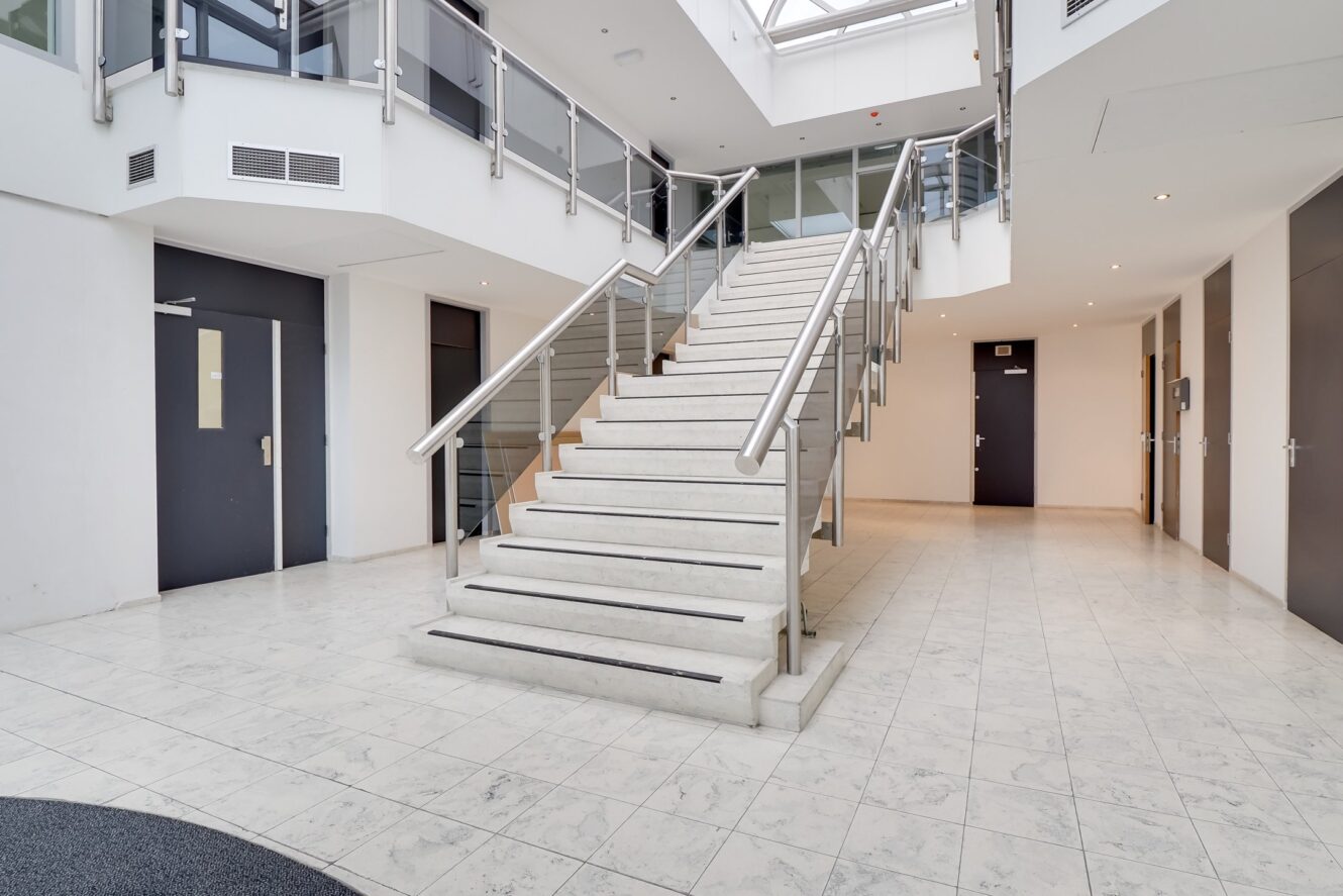 Ruime, moderne lobby van het gebouw met een centrale witte trap, glazen leuningen, tegelvloer en deuren langs de gang. Natuurlijk licht valt binnen door een dakraam erboven.