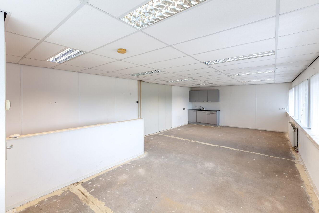 Een leeg kantoor met kale vloeren, witte muren, tl-verlichting aan het plafond en een kleine kitchenette bij de achterwand.