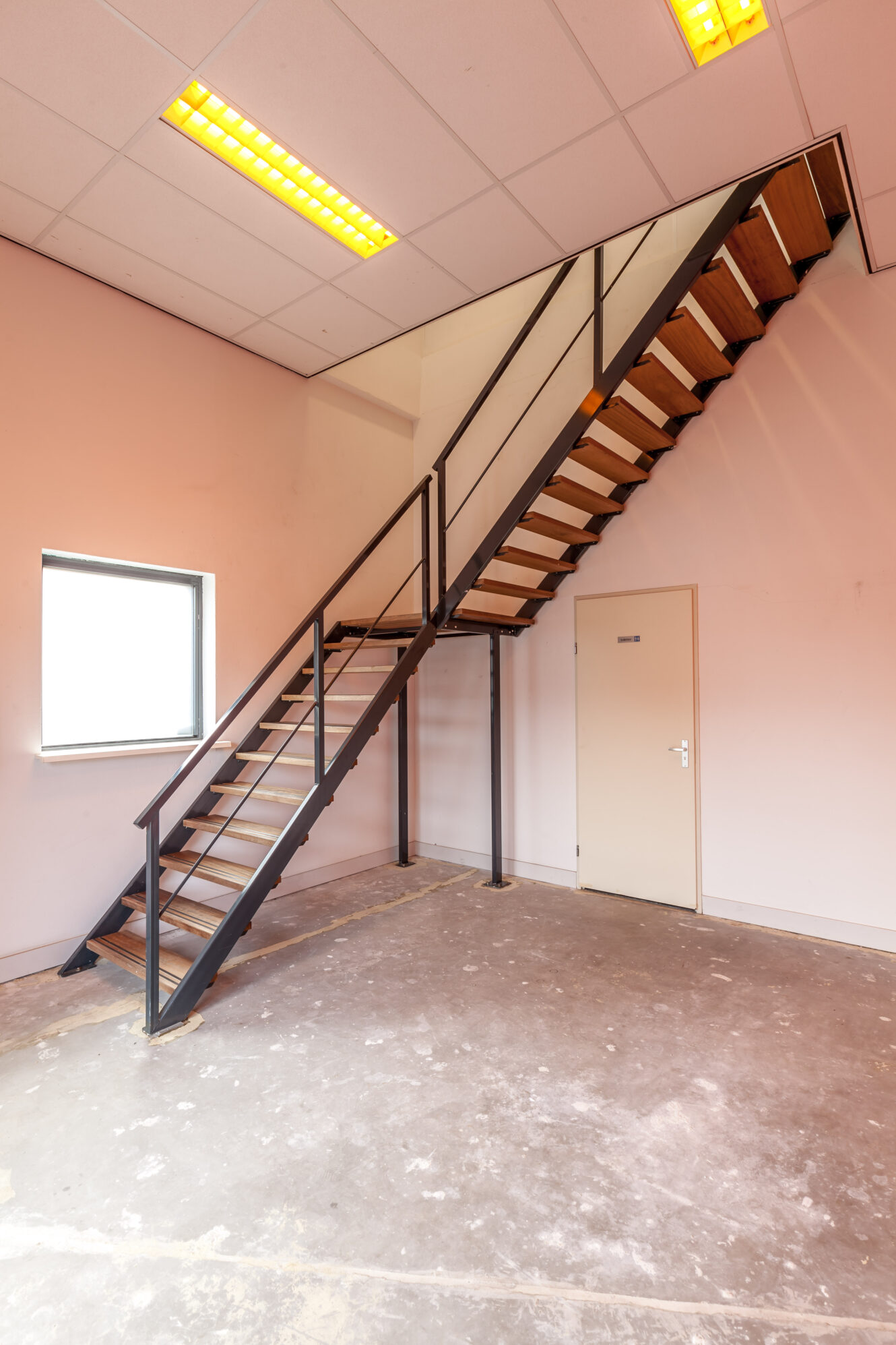 Een metalen trap met houten treden leidt naar een bovenverdieping in een kale kamer met een gesloten deur, een raam en tl-verlichting aan het plafond.