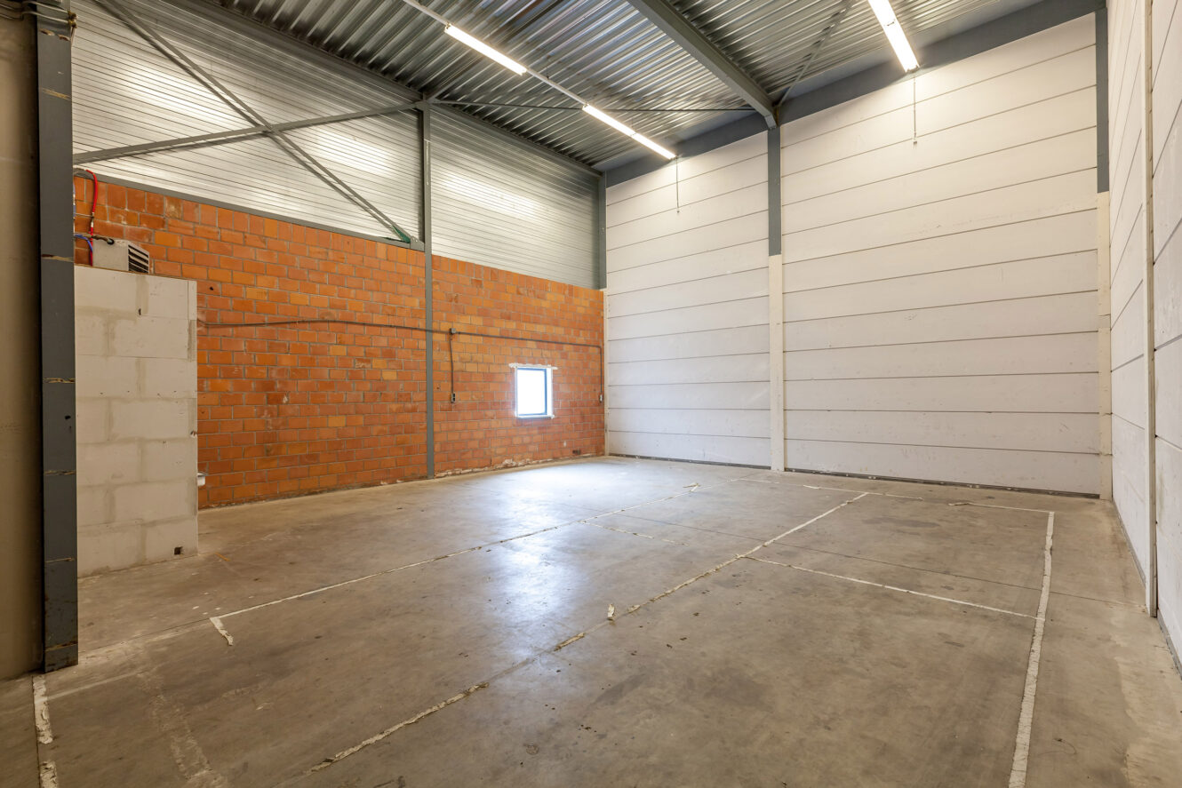 Lege industriële opslagruimte met betonnen vloer, muren van baksteen en witte panelen, hoog plafond, plafondverlichting en een klein raam in één van de muren.