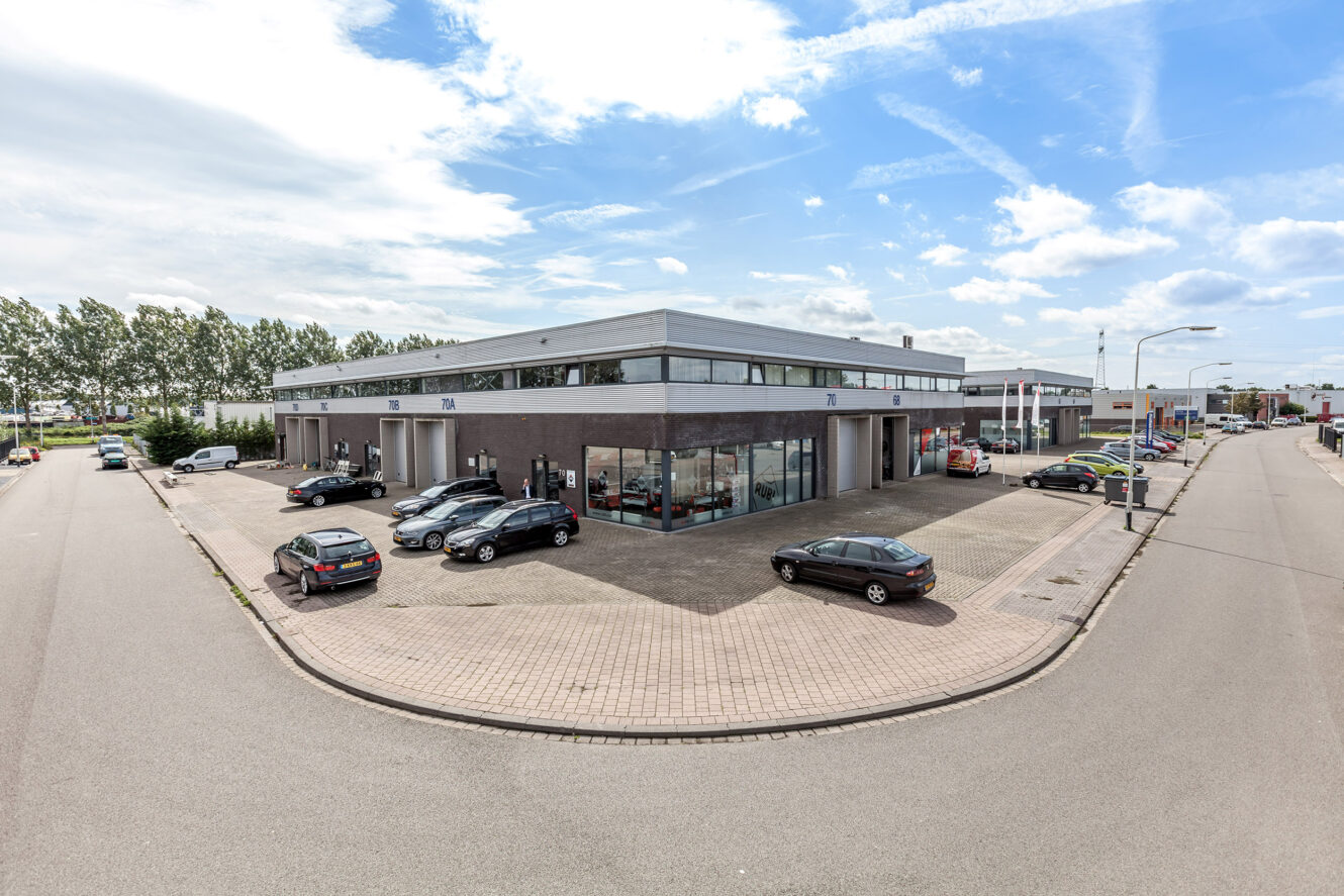 Een modern industrieel gebouw met grote ramen op een straathoek, omringd door geparkeerde auto’s en een grotendeels lege weg onder een gedeeltelijk bewolkte hemel.