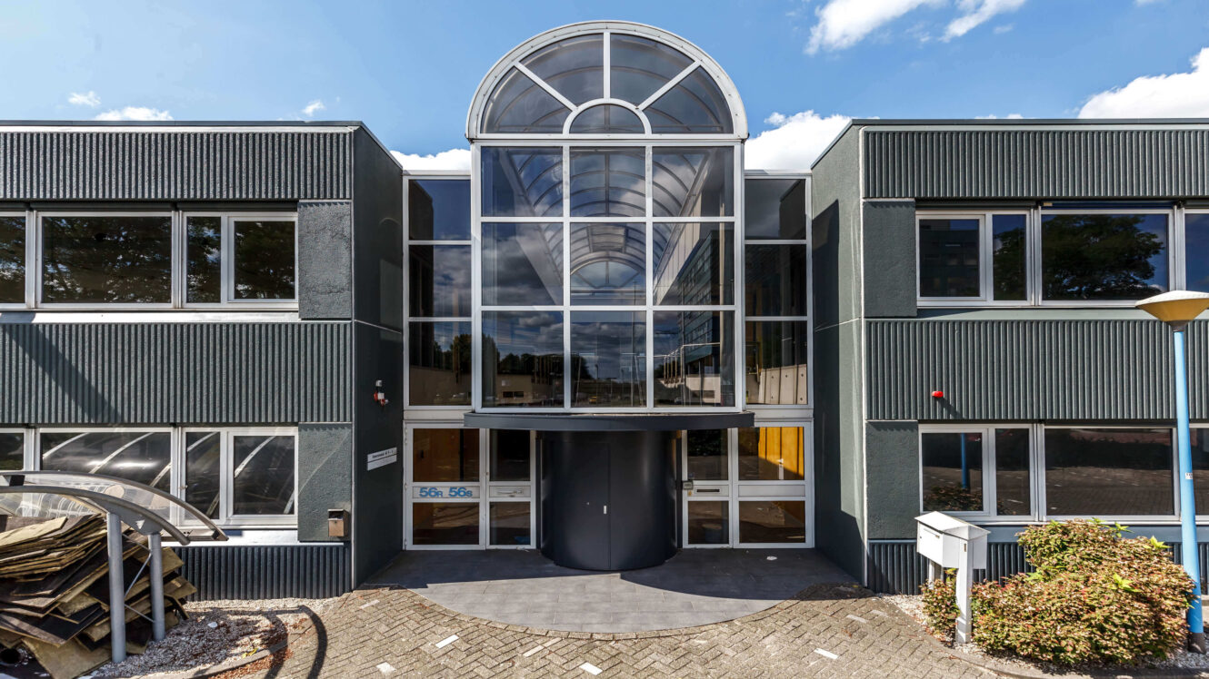 Modern kantoorgebouw van twee verdiepingen met grote glazen ingang, gewelfde luifel en ramen rondom onder een gedeeltelijk bewolkte hemel.