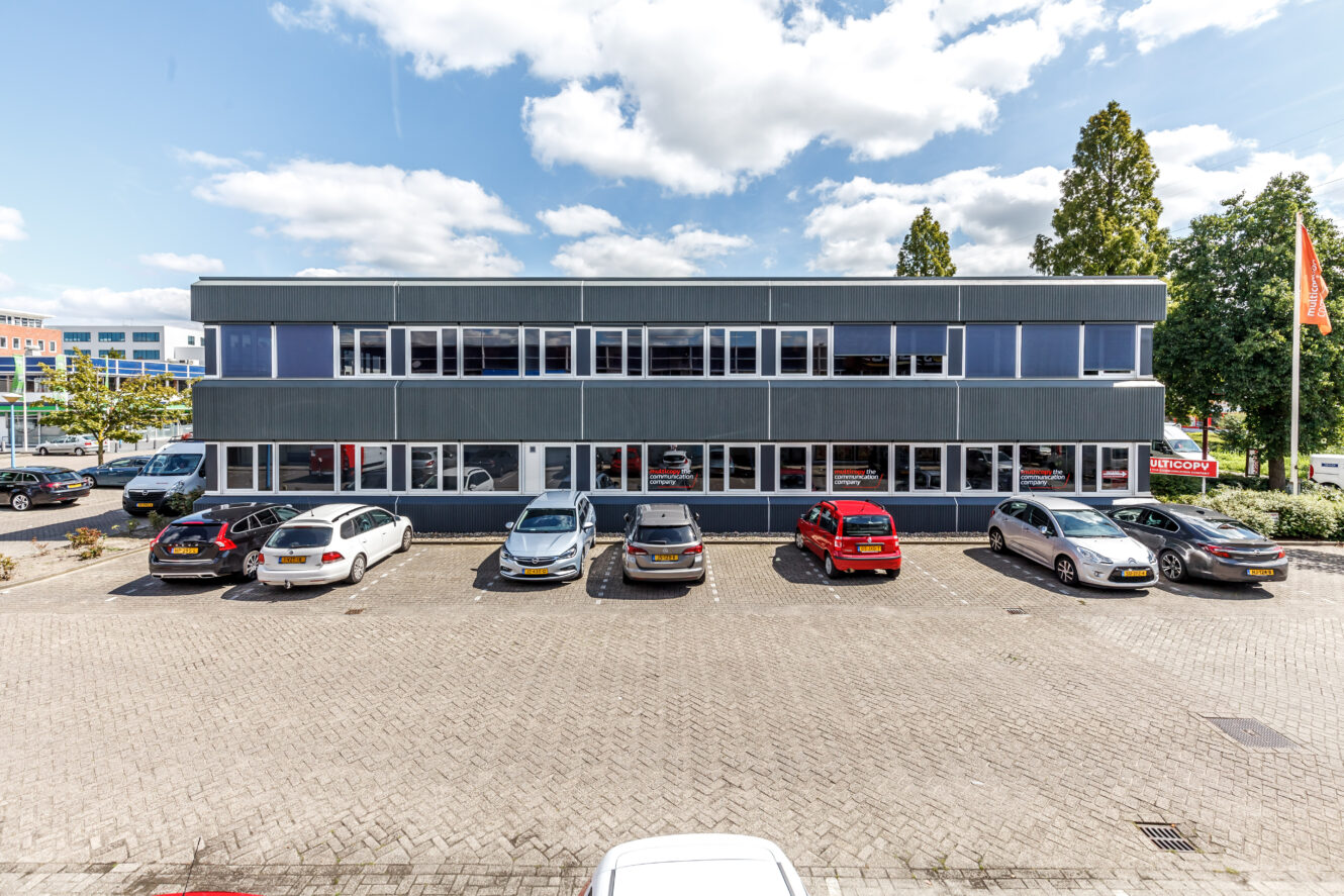 Een kantoorgebouw van twee verdiepingen met grote ramen, een aantal geparkeerde auto's ervoor en een heldere, halfbewolkte lucht erboven.