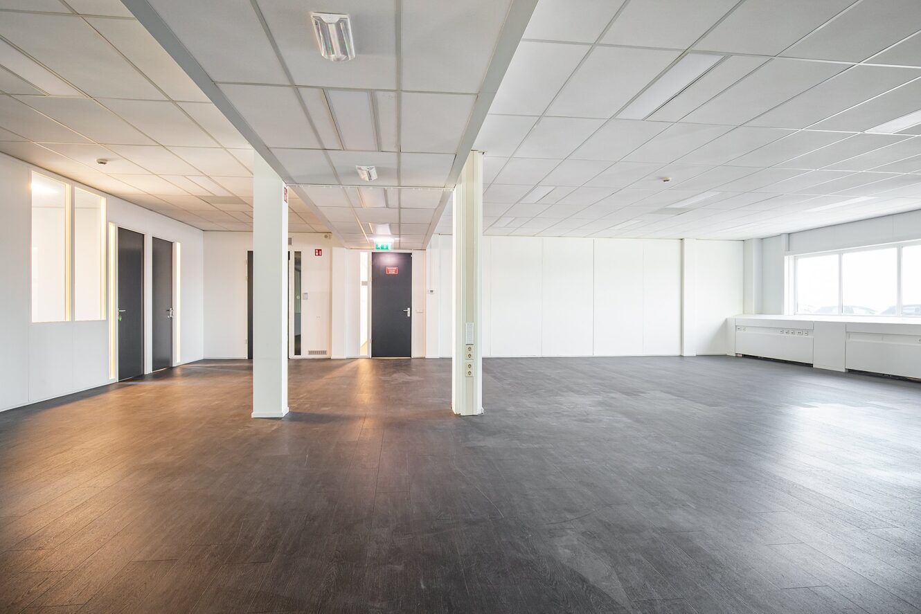 Ruime, lege kantoorkamer met witte muren, grote ramen rechts, zwarte deuren links en een donkere vloer. Het plafond heeft rasterpanelen en ingebouwde verlichting.