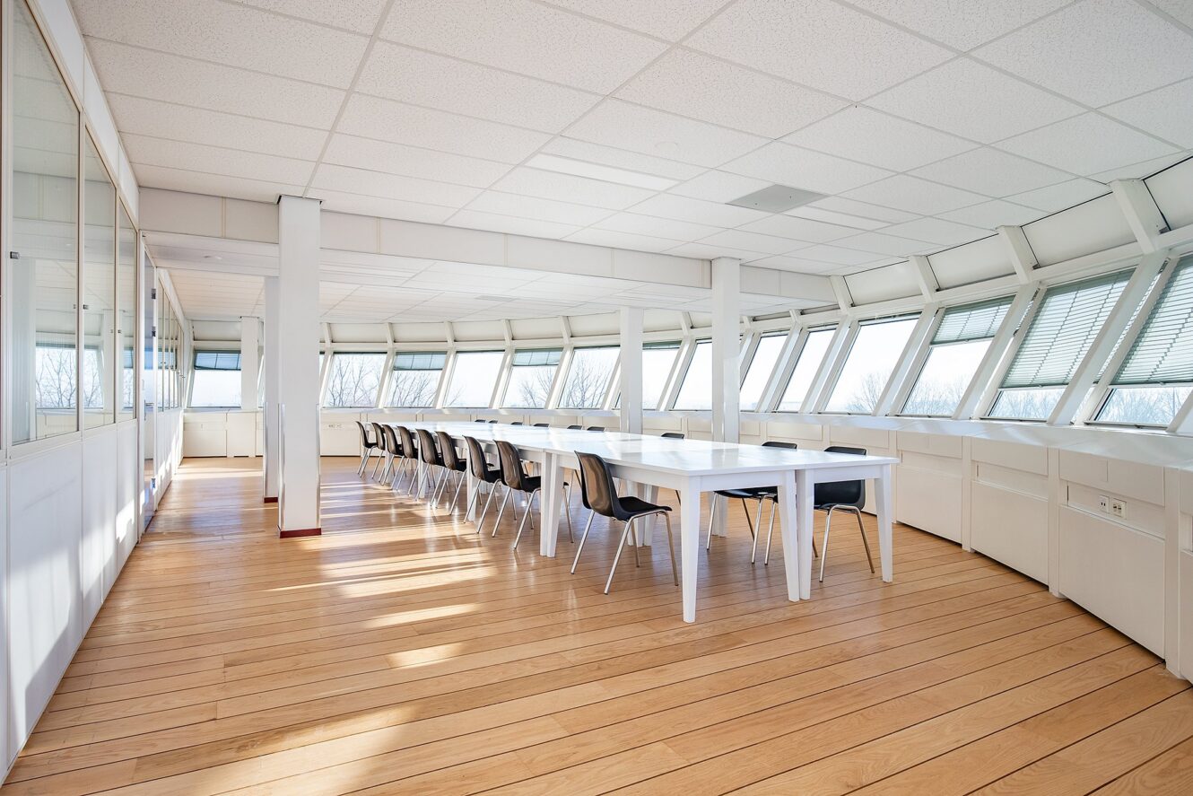 Een lichte, moderne vergaderruimte met lange witte tafels, zwarte stoelen, grote ramen en hardhouten vloeren. De ruimte is leeg en gevuld met natuurlijk licht.