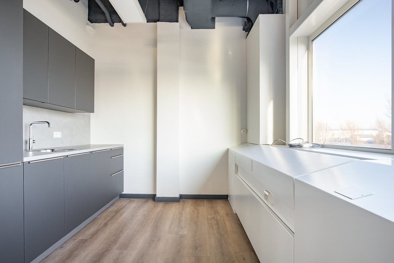 Minimalistische keuken met grijze kastjes, witte werkbladen, roestvrijstalen gootsteen, groot raam en lichte houten vloer.