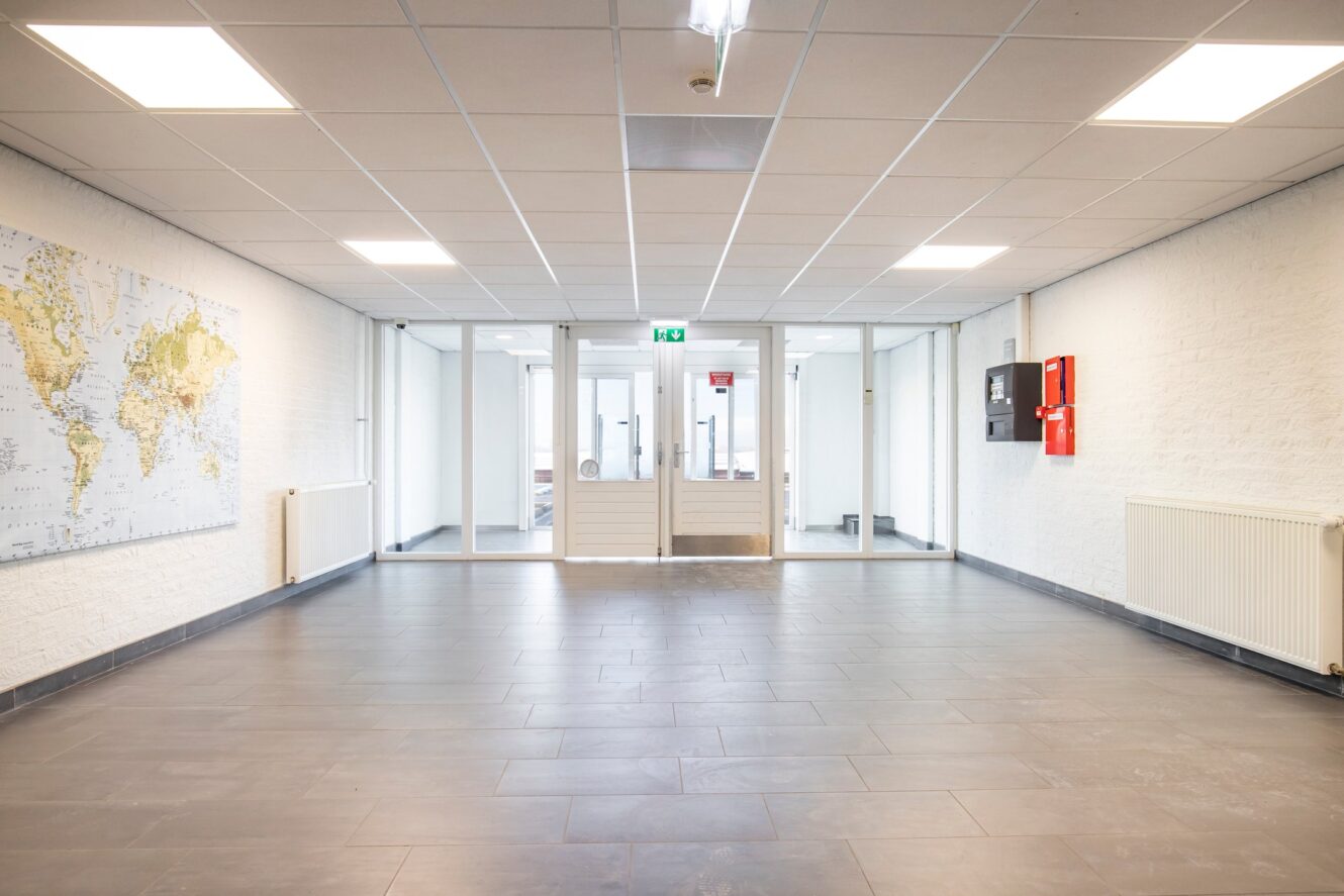 Een schone, lege gang met grijze tegelvloer, witte muren, een wereldkaart aan de linkerkant, brandveiligheidsapparatuur aan de rechterkant en glazen deuren die naar kantoren aan het einde leiden.
