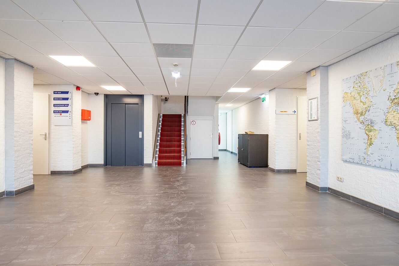 Een schone, lege hal met grijze tegelvloer, een trap met rode vloerbedekking, een lift, richtingaanwijzers, witte muren en een wereldkaart op de rechtermuur.
