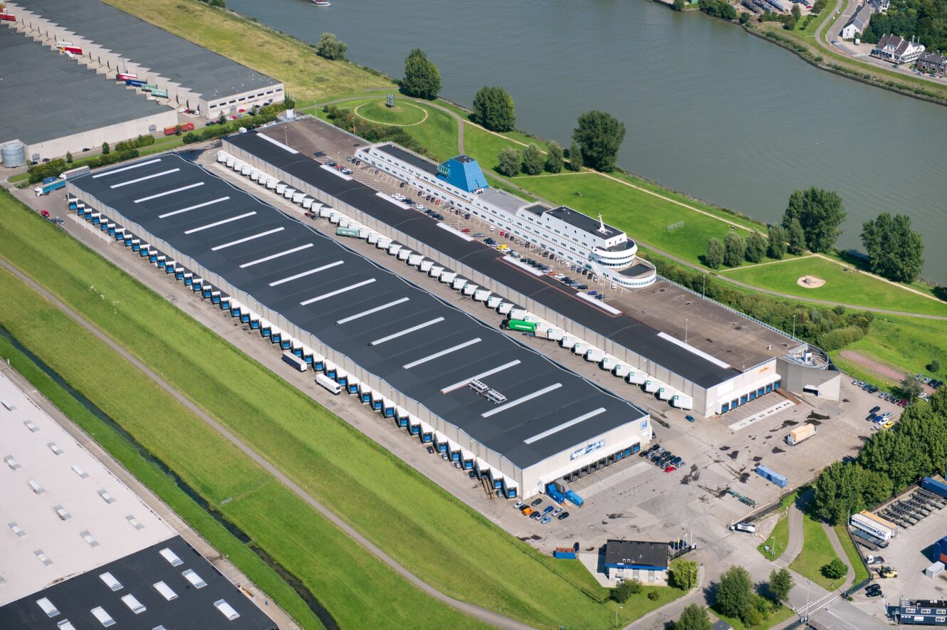 Luchtfoto van een groot industrieel magazijncomplex met laadkades, vrachtwagens, nabijgelegen kantoorgebouwen en een rivier op de achtergrond.