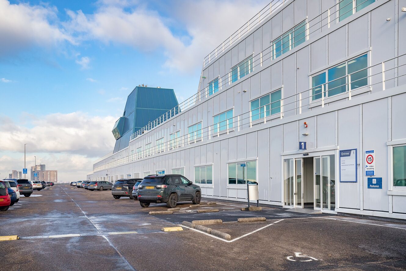 Een modern wit gebouw met grote ramen en verschillende geparkeerde auto's op een verharde parkeerplaats onder een gedeeltelijk bewolkte hemel.