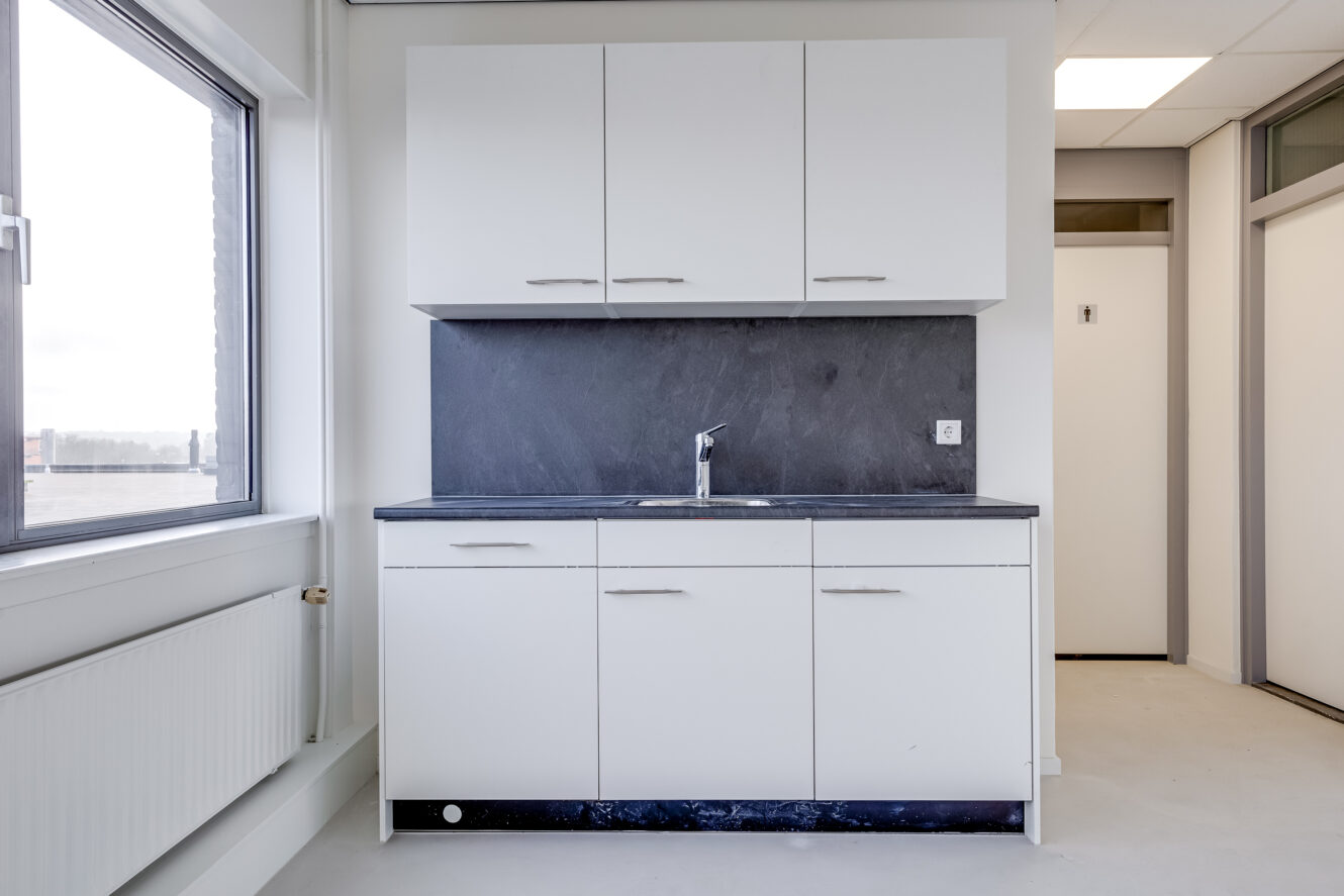 Een klein keukentje met witte kastjes, een donker aanrecht, een kraan aan de muur en een groot raam aan de linkerkant.