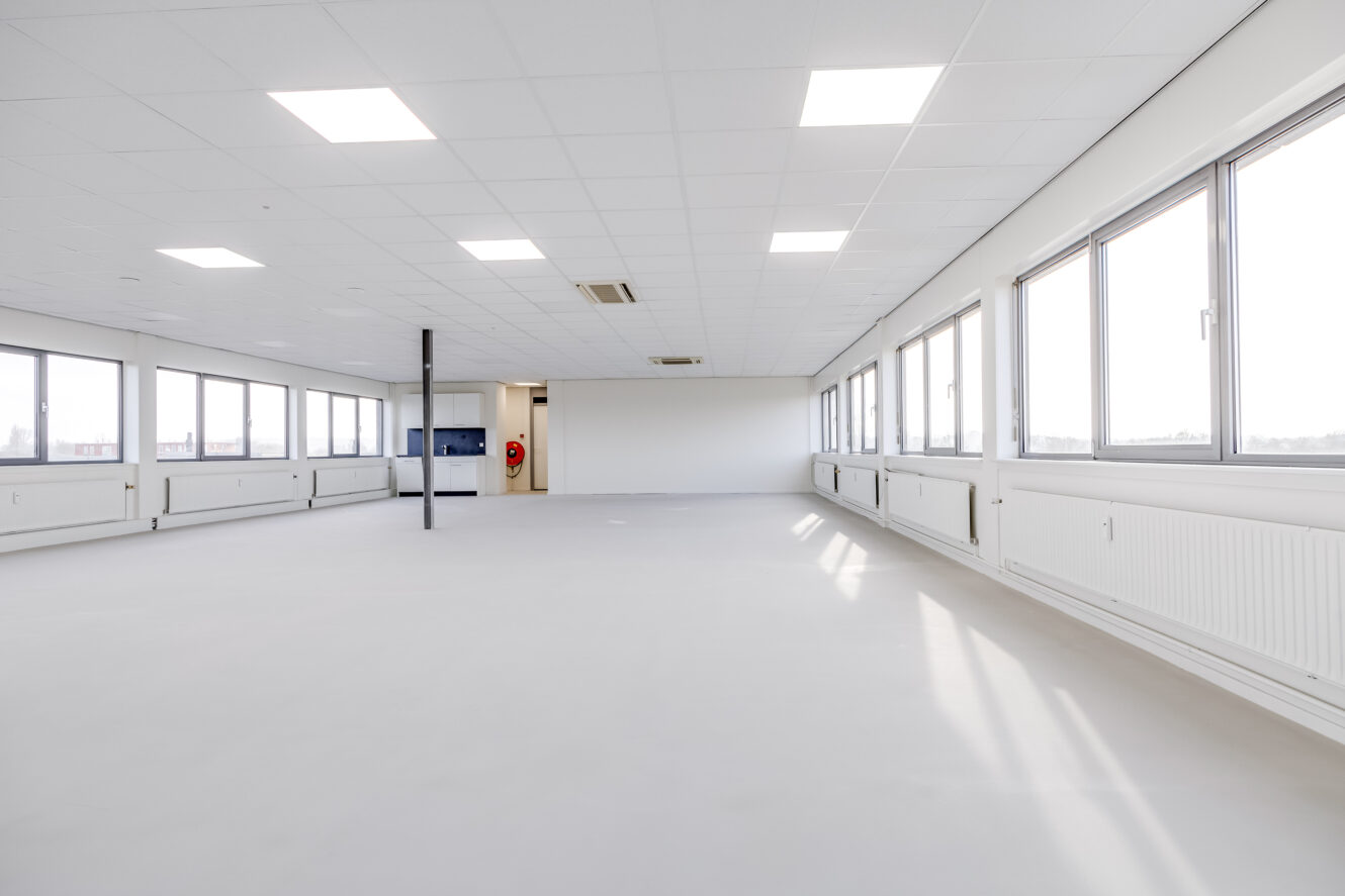 Ruime, lege kantoorkamer met witte muren, heldere verlichting aan het plafond, grote ramen aan één kant en een paal in het midden.