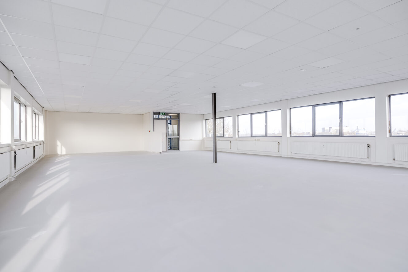 Een grote, lege, witte kantoorruimte met veel ramen, witte muren en een enkele ondersteunende kolom in het midden.