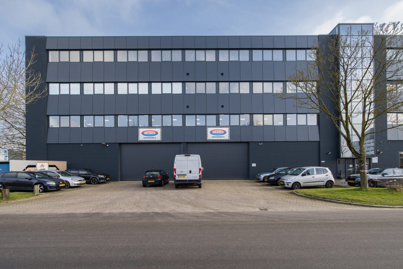 Een modern kantoorgebouw van vier verdiepingen met grote ramen, geparkeerde auto's en een wit busje ervoor. Boven de centrale ingang hangen twee borden.