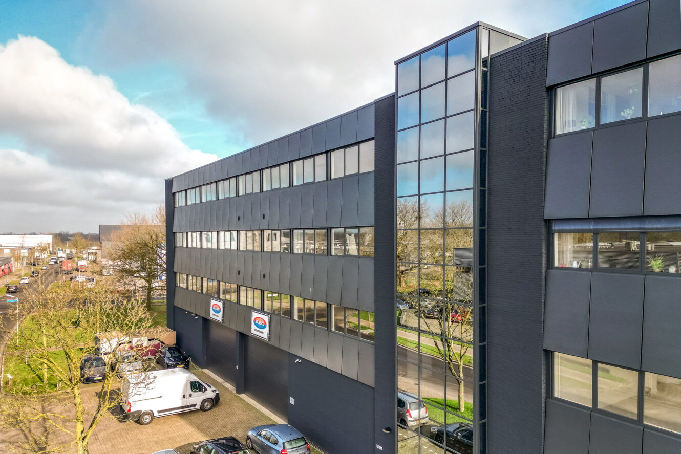 Een modern kantoorgebouw met grote glazen ramen en een aantal geparkeerde voertuigen ervoor, gelegen aan een straat met bomen en een bewolkte lucht.