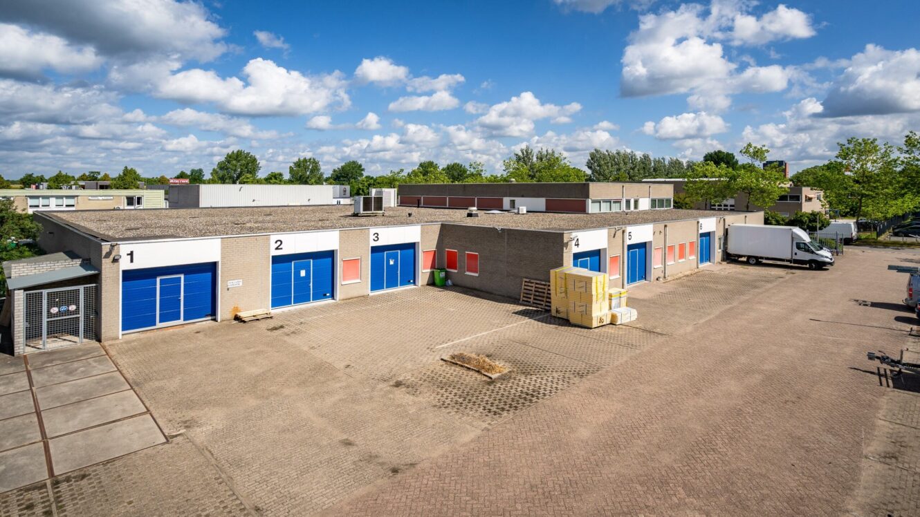 Klein industrieel complex met meerdere opslagunits, genummerde deuren, blauwe garagedeuren, een paar nutsvoorzieningen, een geparkeerde witte vrachtwagen en een gedeeltelijk bewolkte lucht boven je hoofd.