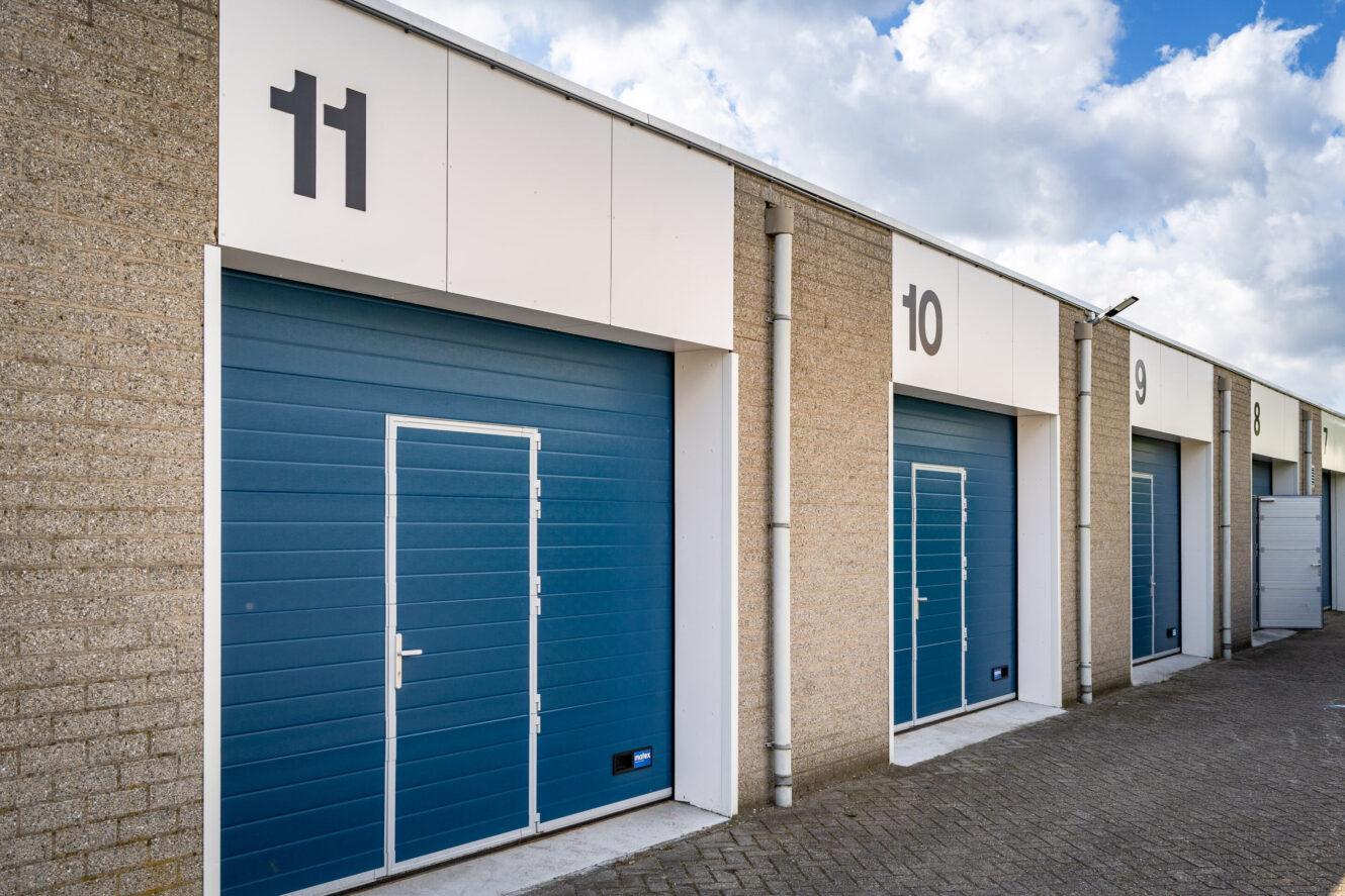 Een rij opslagdeuren genummerd van 9 tot 11, elk met een blauwe roldeur en een kleinere ingebouwde toegangsdeur, tegen een bakstenen en witte gevel onder een gedeeltelijk bewolkte hemel.