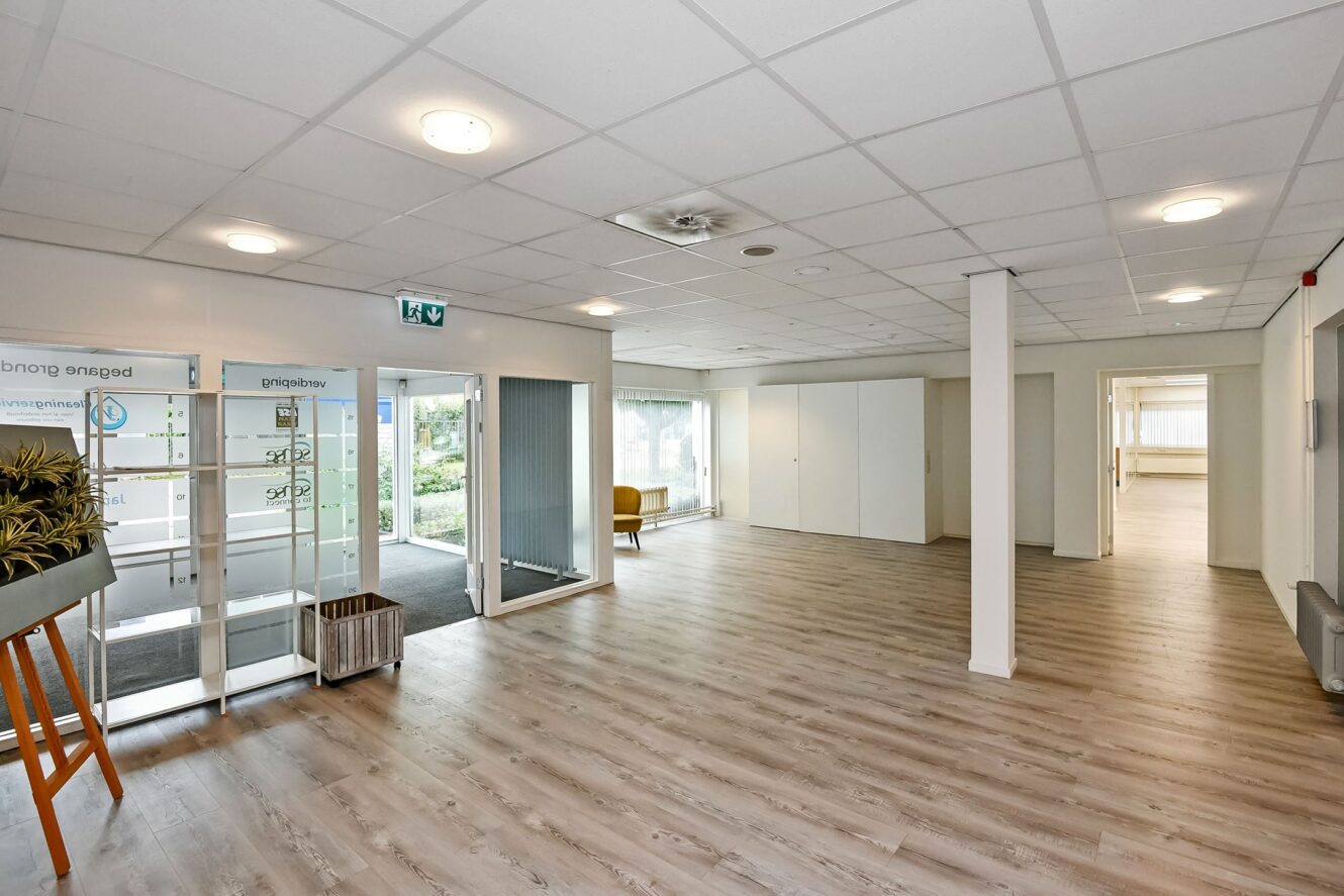 Ruime, moderne lobby met lichte houten vloeren, witte muren, plafondverlichting, glazen toegangsdeuren en grote ramen die voor natuurlijk licht zorgen.