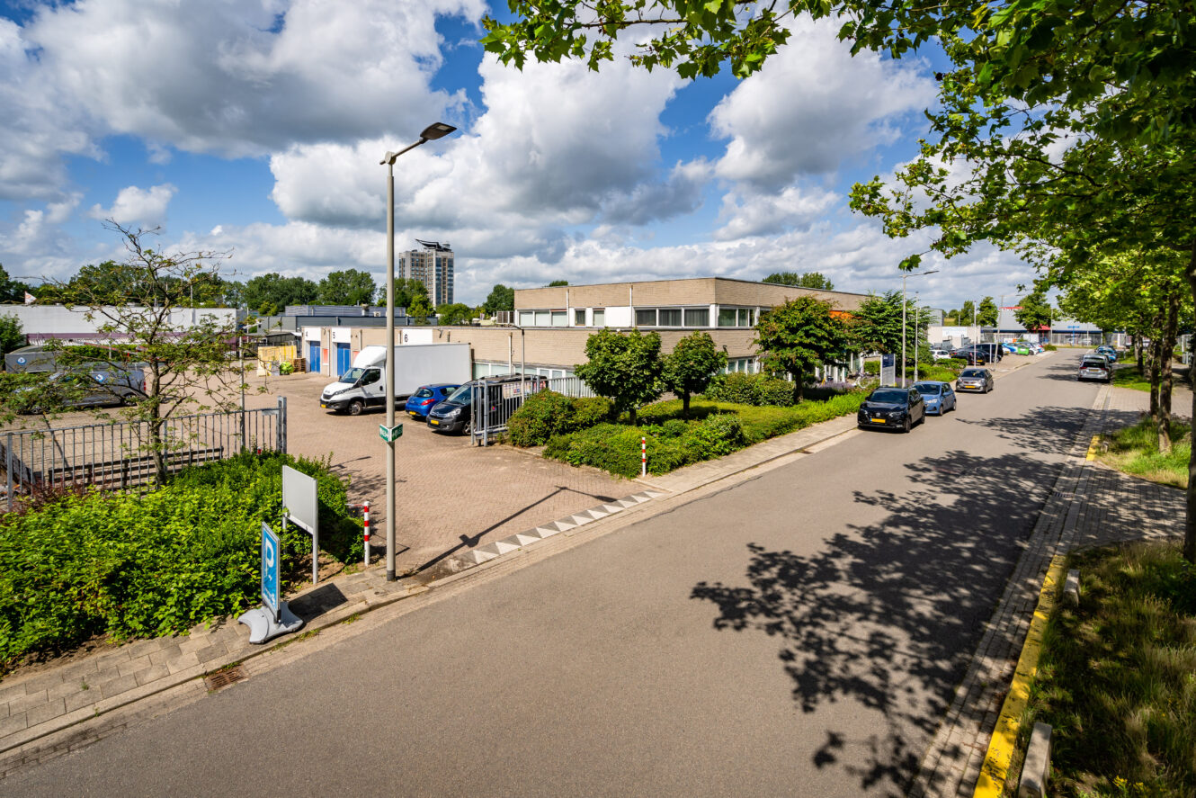 Een rustige straat met geparkeerde auto's en bomen, grenzend aan een laag industrieel of kantoorgebouw onder een gedeeltelijk bewolkte hemel.