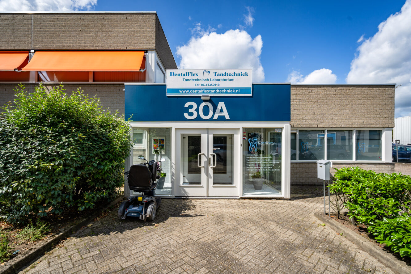 Een ingang van een tandartspraktijk op 30A met glazen deuren, een bord erboven, een scootmobiel die buiten geparkeerd staat en groen aan beide kanten van het pad.