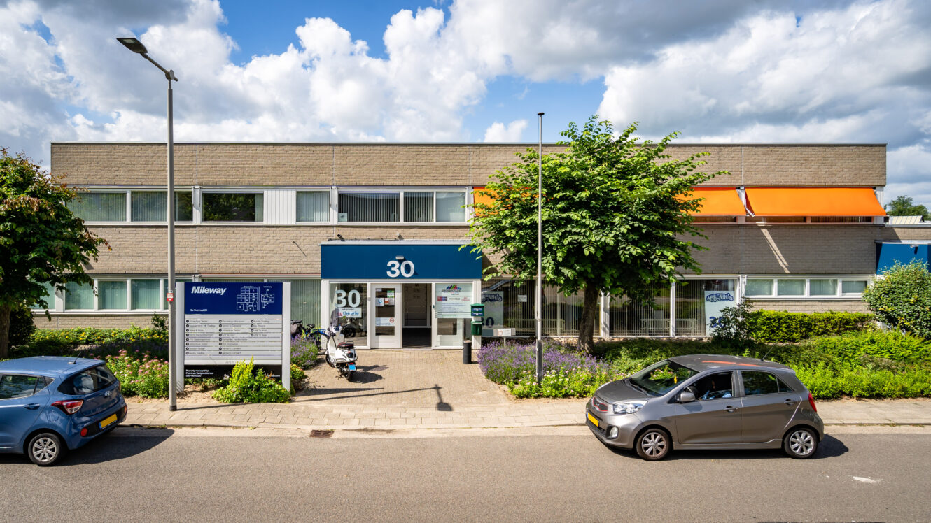 Een kantoorgebouw van twee verdiepingen met het nummer 30 boven de ingang, geparkeerde auto's ervoor, een bord met bedrijven en bomen en struiken langs het trottoir.