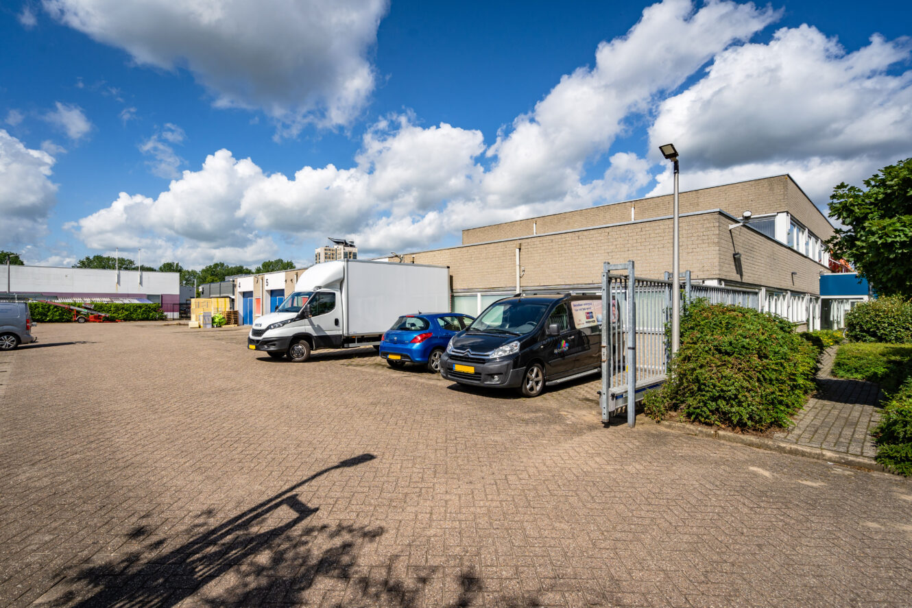 Een verharde parkeerplaats met verschillende geparkeerde auto's en bestelwagens naast een beige industrieel gebouw onder een gedeeltelijk bewolkte hemel.