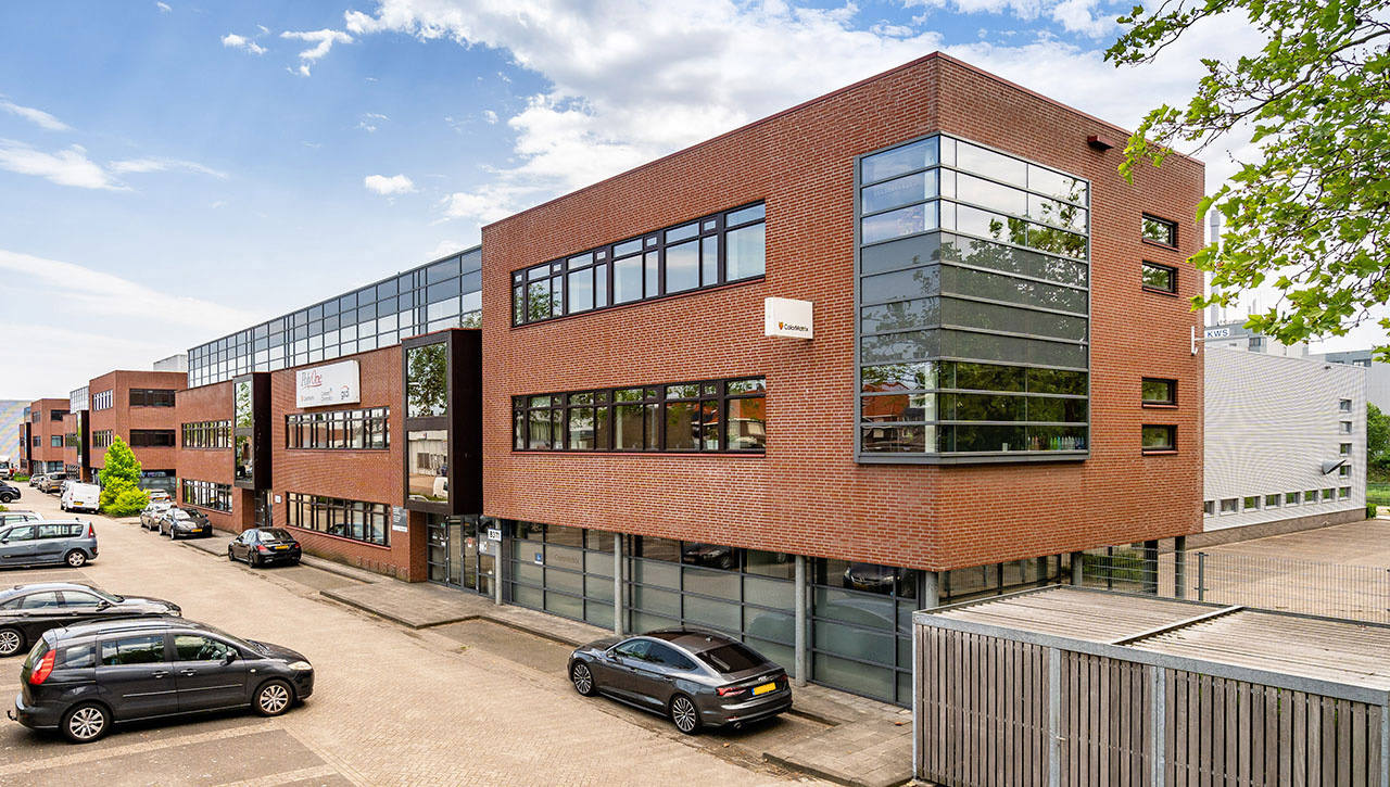 Modern bakstenen kantoorgebouw van twee verdiepingen met grote glazen ramen, een aantal geparkeerde auto's ervoor en een strakblauwe lucht met een paar wolken.