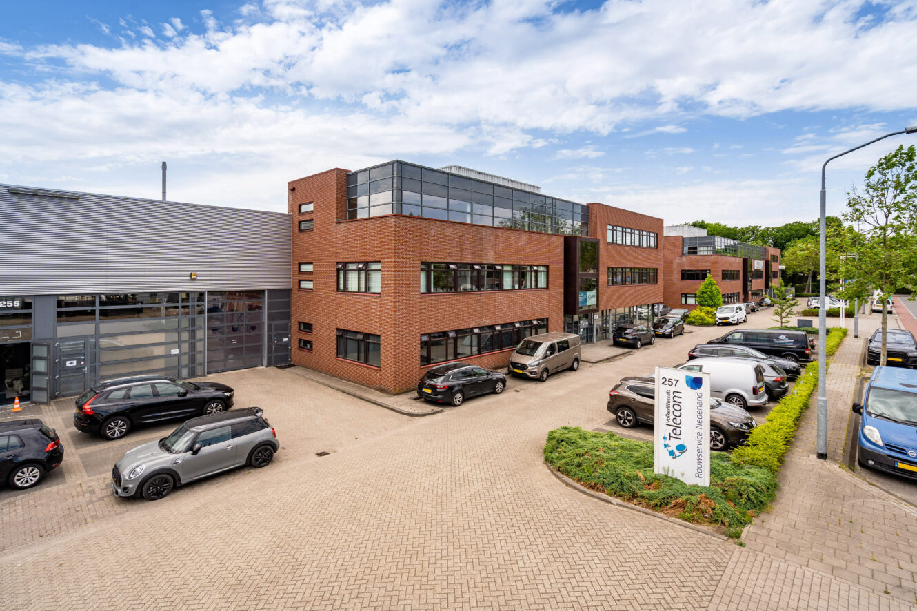 Modern kantoorgebouw met grote ramen, bakstenen en metalen buitenkant en een parkeerplaats met meerdere auto's; op een bord aan de voorkant staat de naam "Telecom" en het straatnummer 257.