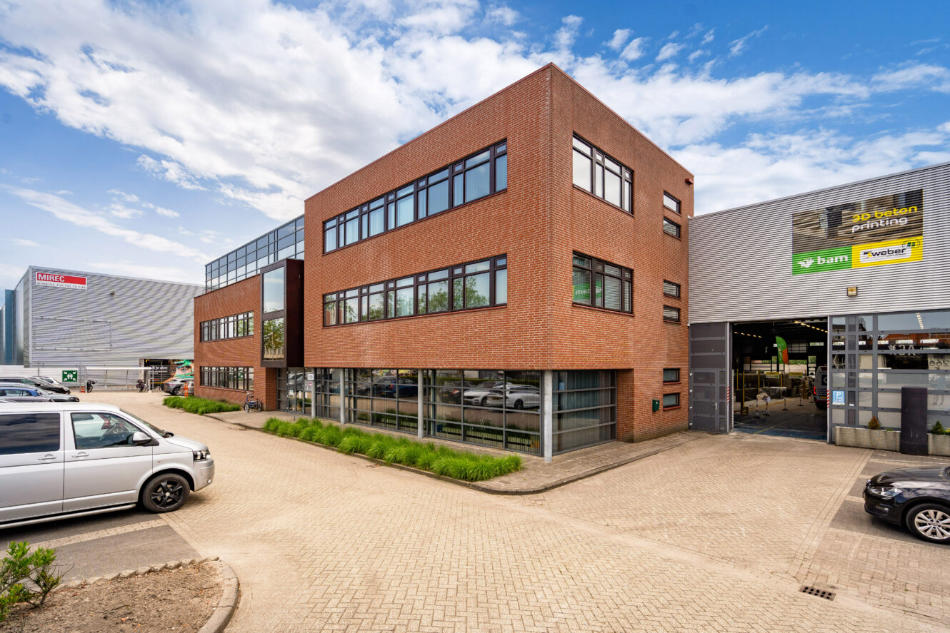 Een modern industrieel kantoorgebouw met grote ramen en een bakstenen gevel, grenzend aan een magazijn; verschillende auto's staan buiten geparkeerd onder een gedeeltelijk bewolkte hemel.