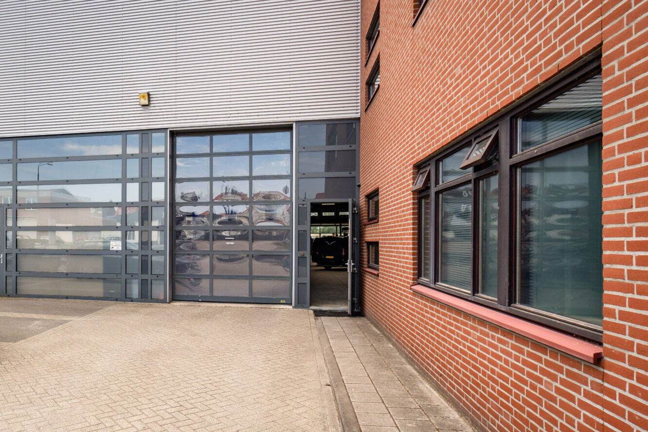 Een magazijningang met een grote glazen garagedeur die gedeeltelijk openstaat en een rode bakstenen buitenmuur met twee ramen aan de rechterkant.