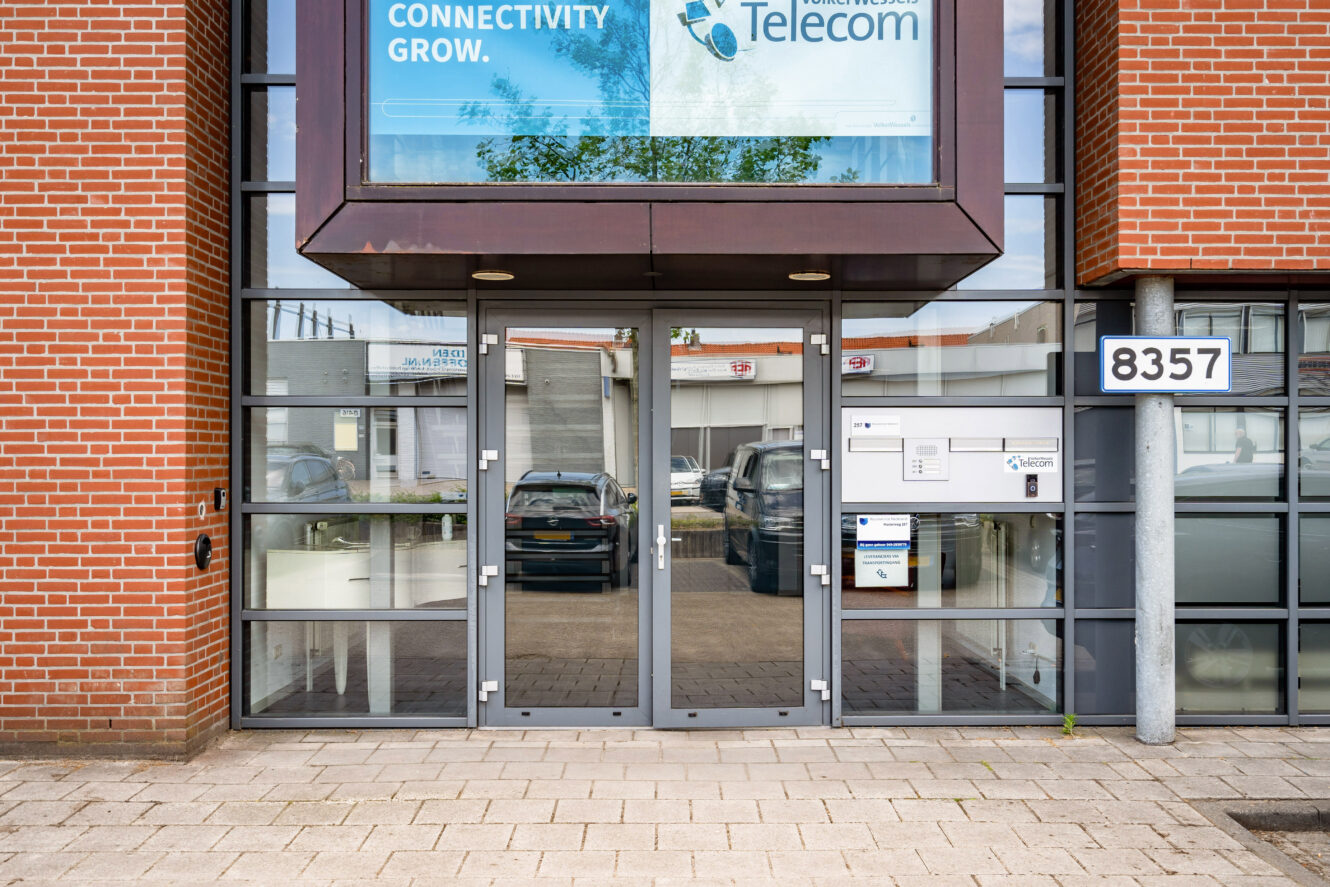 Glazen toegangsdeuren van een commercieel gebouw met een bord 8357 naast de deur, brievenbussen en een groot spandoek voor een telecombedrijf erboven. Auto's zijn zichtbaar door het glas.