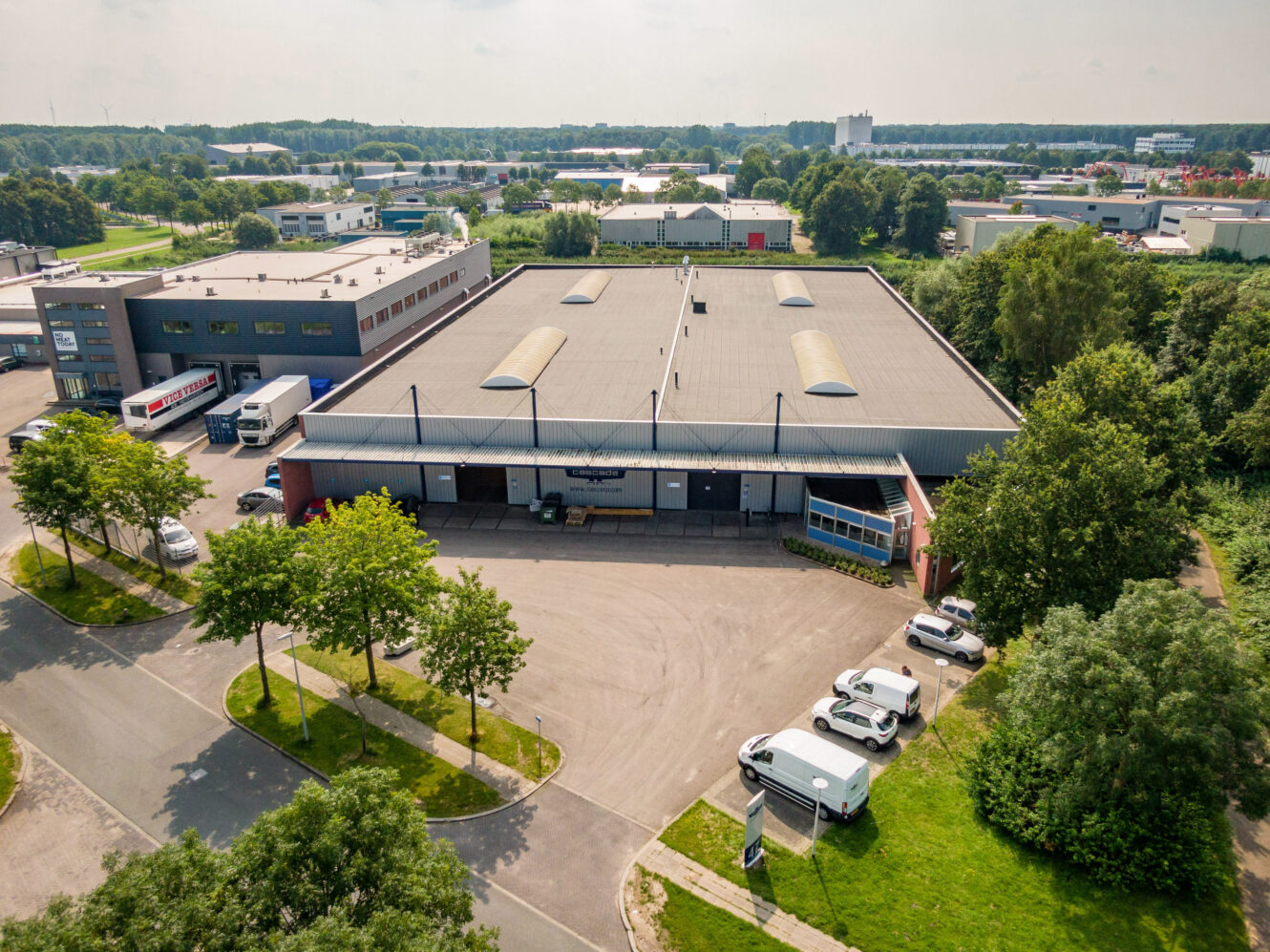 Luchtfoto van een groot industrieel magazijn met laadkades, vrachtwagens en geparkeerde voertuigen, omringd door bomen en andere industriële gebouwen.