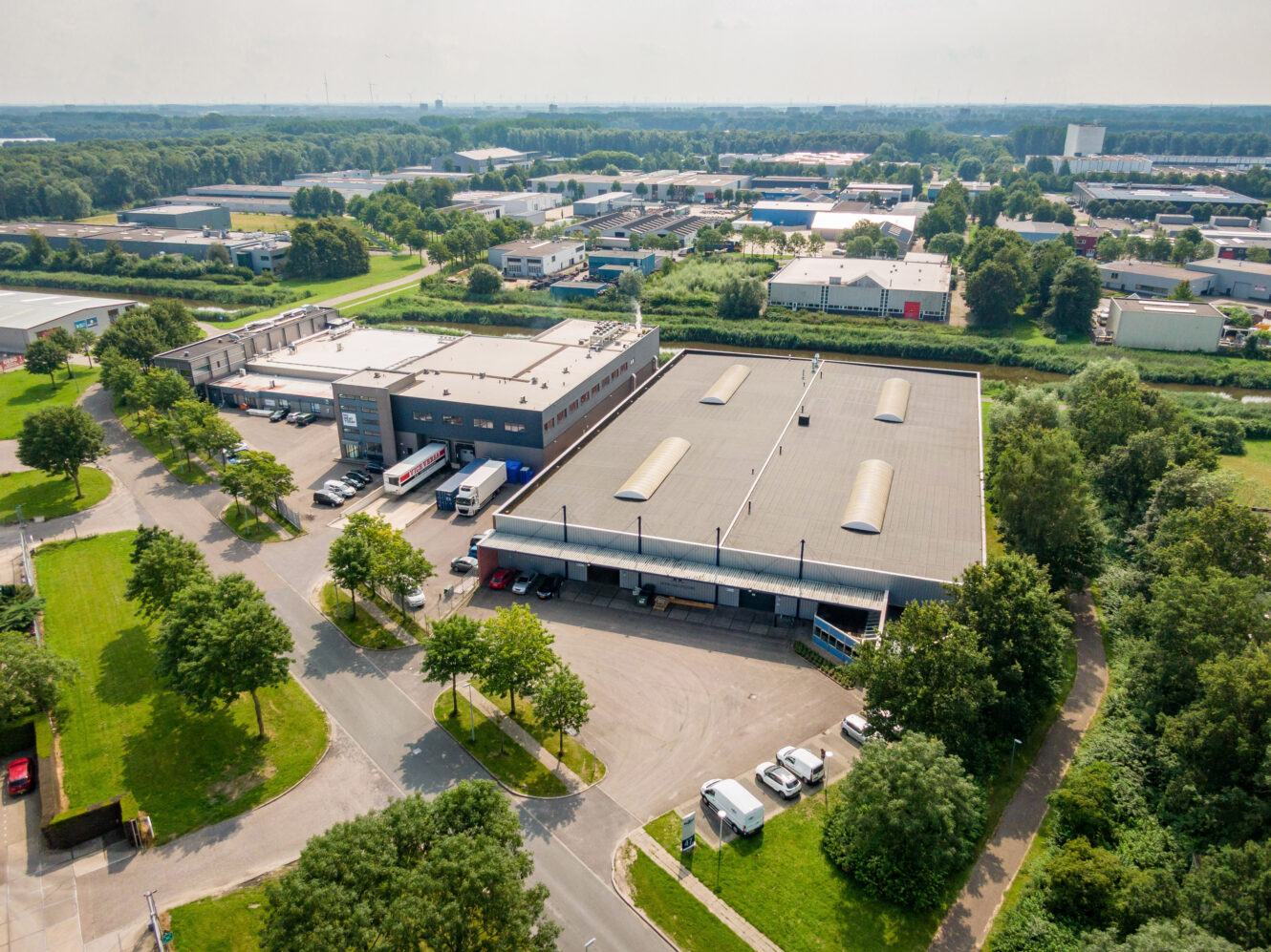 Luchtfoto van een industrieel gebied met grote magazijngebouwen, vrachtwagens geparkeerd bij laadkades en omringend groen.