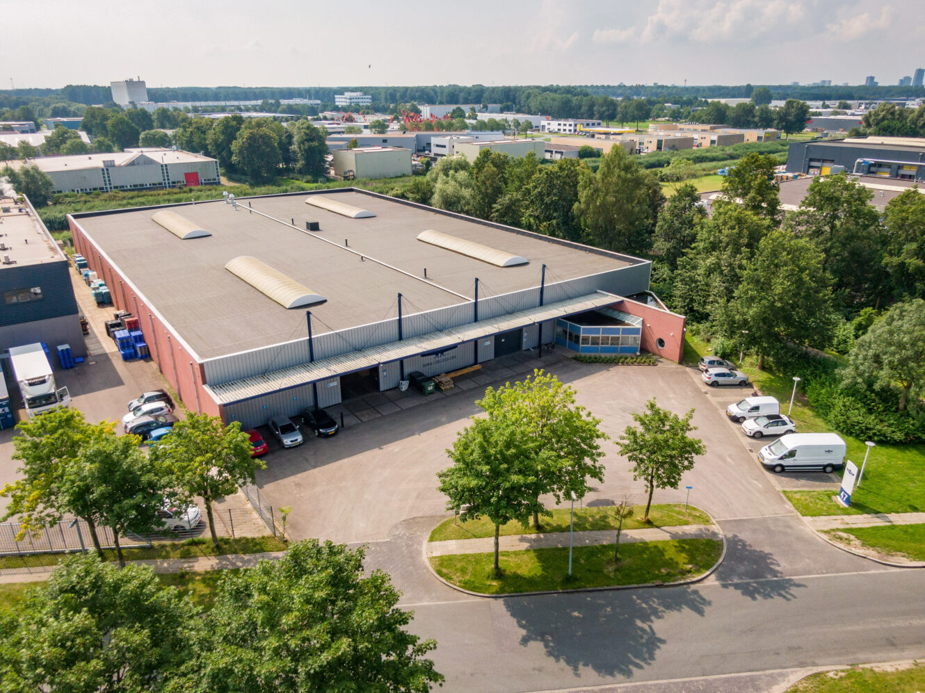 Luchtfoto van een groot industrieel magazijn met laadkades, verschillende geparkeerde vrachtwagens en auto's, omringd door bomen en andere industriële gebouwen.