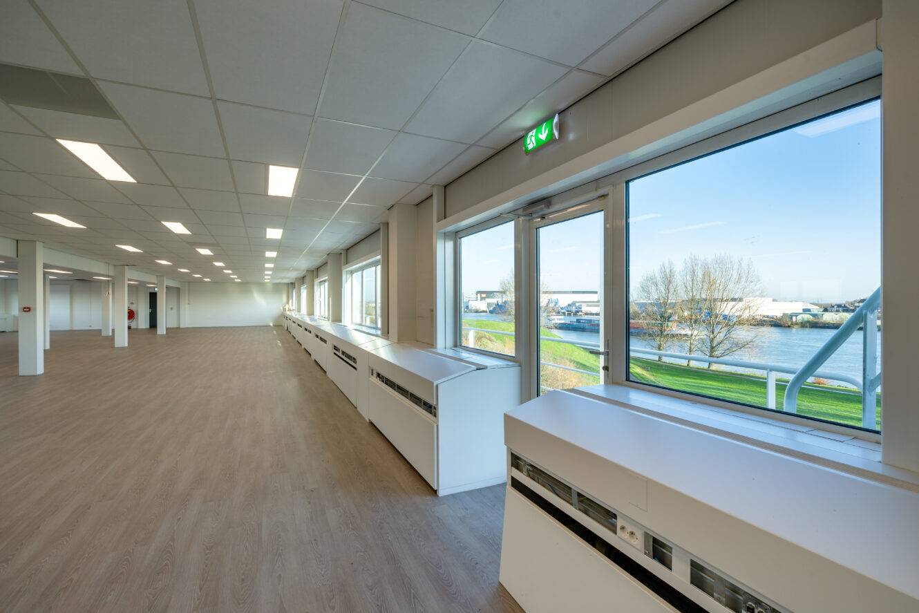 Ruim, leeg kantoor met grote ramen, houten vloeren, witte muren en uitzicht op een rivier en gebouwen buiten.