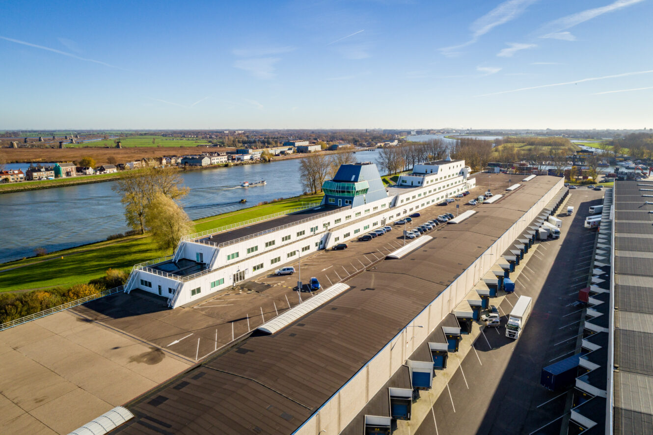 Een groot, wit gebouw in de vorm van een schip ligt bij een rivier, met parkeerplaatsen en vrachtwagens ernaast op een heldere, zonnige dag.