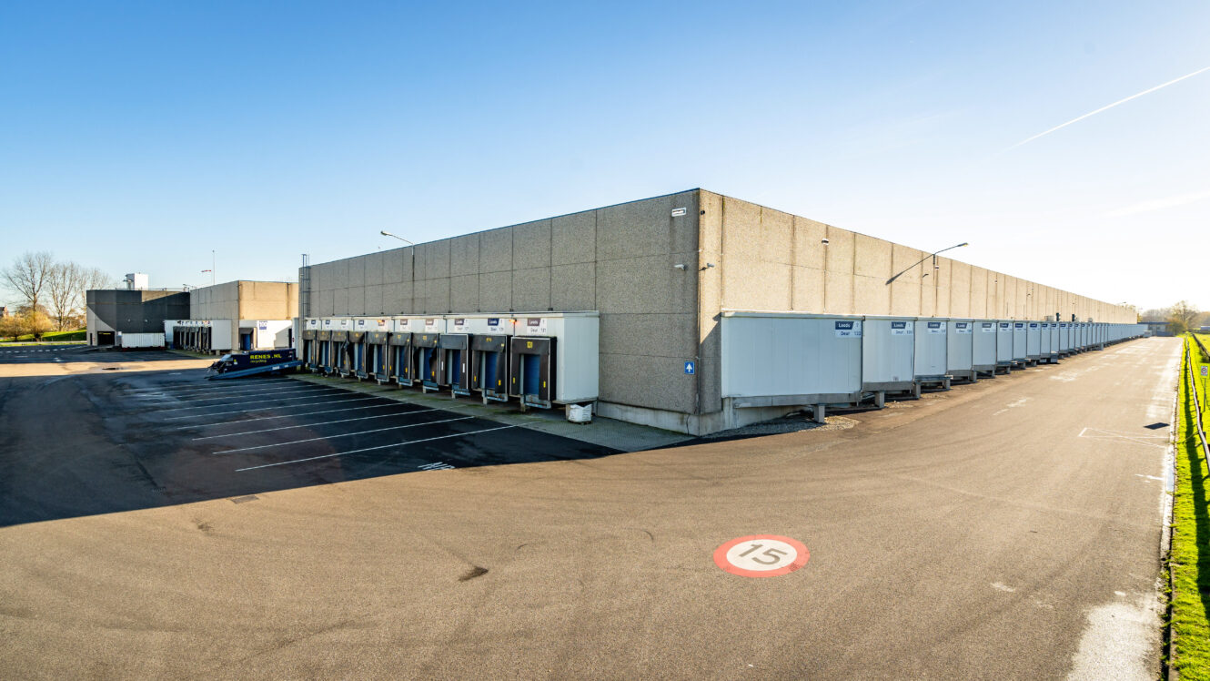 Groot industrieel magazijn met meerdere laadkades en geparkeerde trailers, onder een strakblauwe hemel, grenzend aan een leeg parkeerterrein en een rijbaan gemarkeerd met een snelheidslimiet van 15.