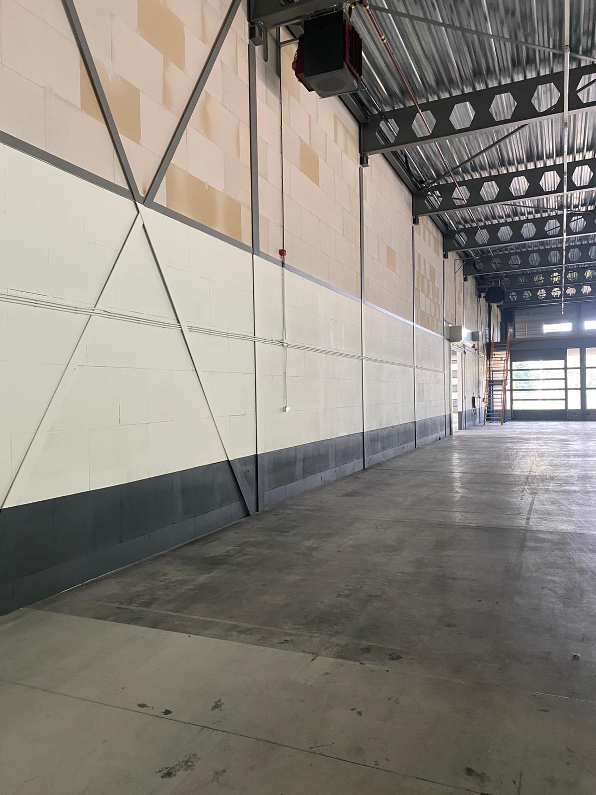 Interieur van een leeg pakhuis met hoge plafonds, zichtbare balken, industriële verlichting en een trap naar een hoger niveau op de achtergrond.
