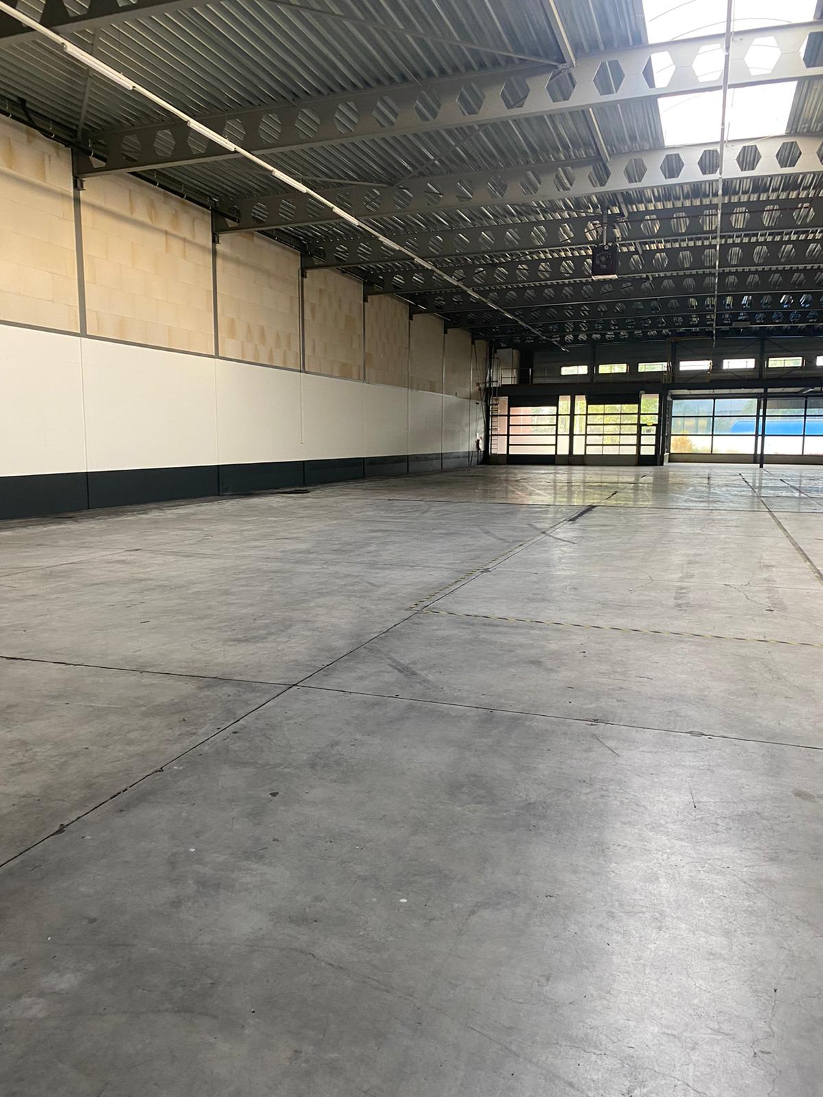 Interieur van een leeg, ruim magazijn met een betonnen vloer, hoog plafond en grote ramen die daglicht binnenlaten.