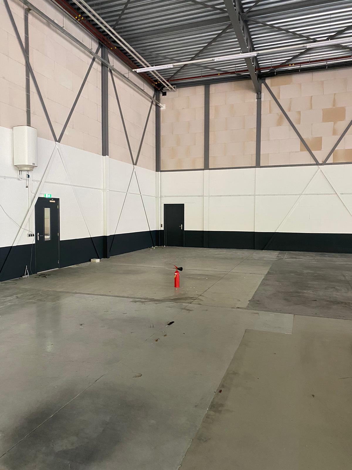 Een leeg industrieel pakhuis met betonnen vloeren, hoge plafonds en twee gesloten deuren; een rode brandblusser staat in het midden van de ruimte.
