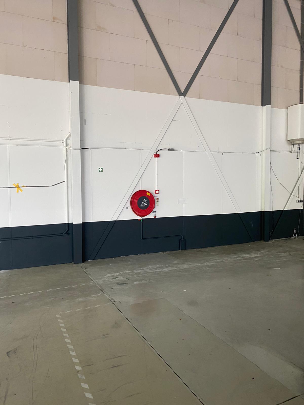 Een overdekte industriële ruimte met een brandslanghaspel aan een witte en grijze muur; de vloer is leeg en lijkt schoon.