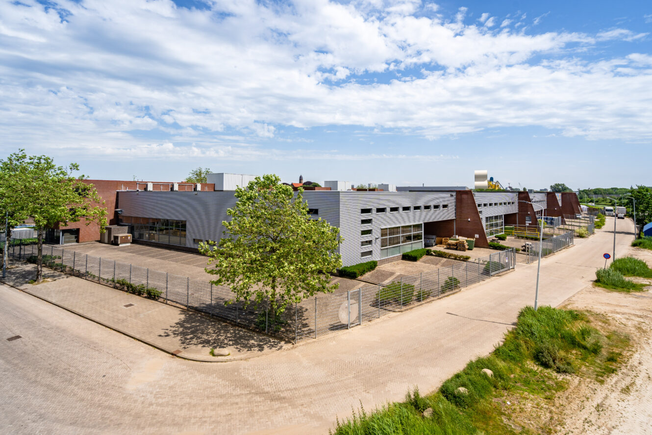 Een modern industrieel gebouw met een omheind terrein, omringd door bomen en verharde wegen onder een gedeeltelijk bewolkte hemel.